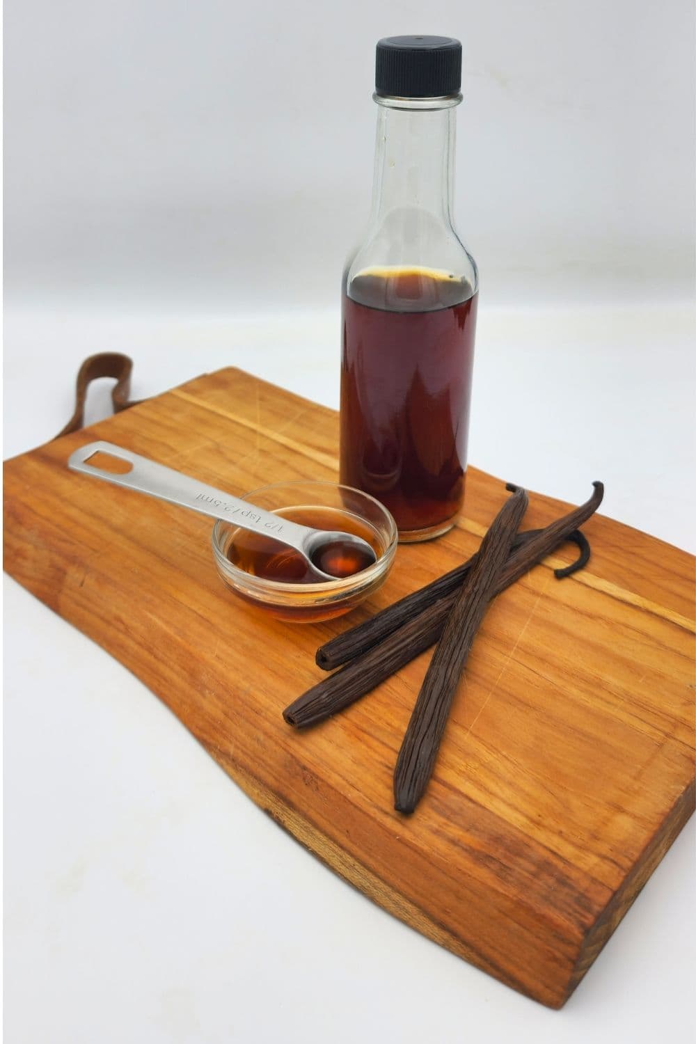 Woozy bottle of deep amber vanilla extract with measuring dish and vanilla beans